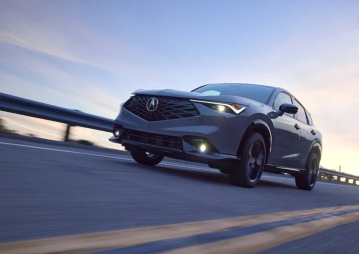 Gray Acura SUV driving along a highway at dusk with its lights on