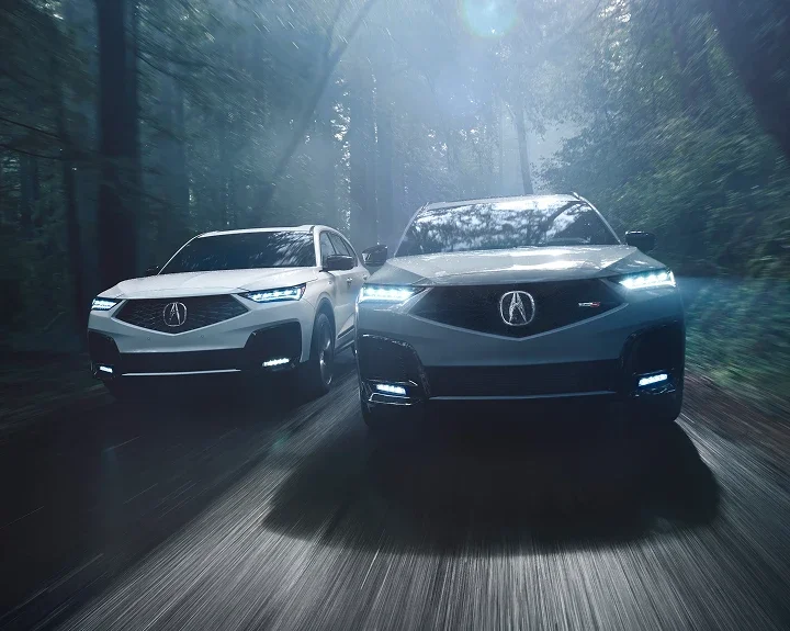 Two white Acura MDX SUVs with headlights on, driving through a misty forest road